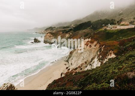 Hoher Winkel der wunderbaren Landschaft des stürmischen Ozeans Wasser mit Schaumwellen waschen den Sandstrand gegen die neblige Hochlandküste unter Grauer Himmel in Big Sur Stockfoto