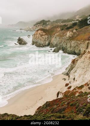 Hoher Winkel der wunderbaren Landschaft des stürmischen Ozeans Wasser mit Schaumwellen waschen den Sandstrand gegen die neblige Hochlandküste unter Grauer Himmel in Big Sur Stockfoto