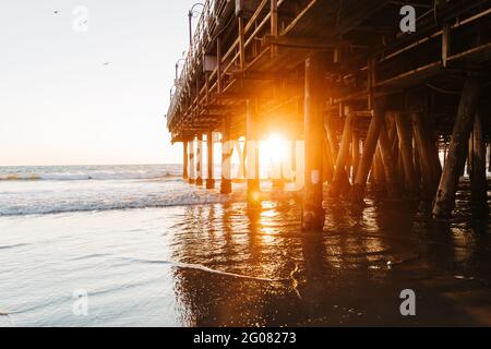 Glühendes Sonnenlicht durchdringt abends die Berge von Santa Monica pier mit friedlichen Wellen am Strand in Kalifornien Stockfoto