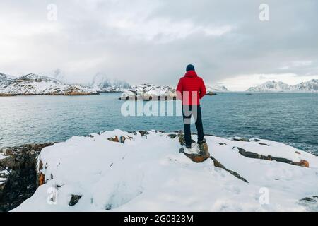 Rückansicht eines anonymen Mannes in Oberbekleidung, der auf einer verschneiten Klippe steht und am Wintertag das plätschernde Meer und den wolkenlosen Himmel auf den Lofoten-Inseln, Norwegen, bewundert Stockfoto