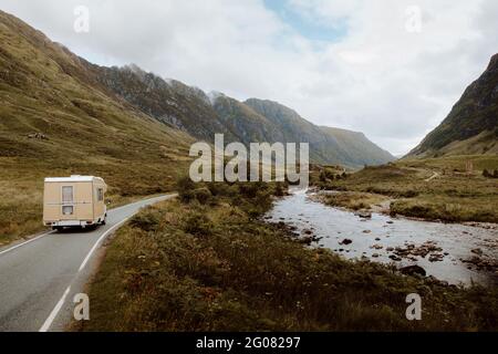 Wohnwagen auf Asphaltstraße in der Nähe von Bach inmitten von Hügeln während Ausflug durch die Landschaft Großbritanniens am bewölkten Tag Stockfoto