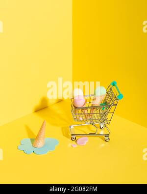 Von oben Zusammensetzung von Miniatur-Shopping-Trolley mit verschiedenen bunten Eiszapfen, die in der Nähe von heruntergefallener, schmelzender blauer Eiscreme platziert werden Auf gelber BA Stockfoto