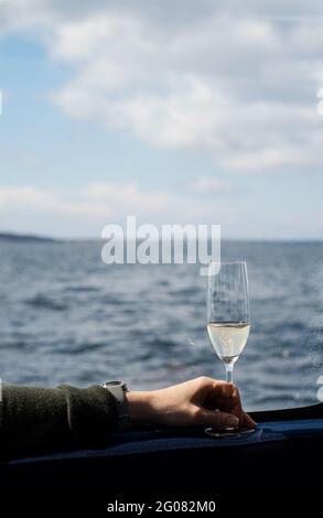 Person, die ein Glas Champagner in der Hand hält, während sie auf einem Boot unterwegs ist. Foto aus dem schwedischen Schärengarten. Stockfoto