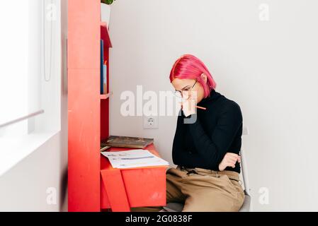 Junge, nachdenkliche, kreative Designerin mit rosafarbenem Haar in Casual Outfit und Brille zeichnen Skizzen, während sie am Schreibtisch arbeiten Zu Hause Stockfoto