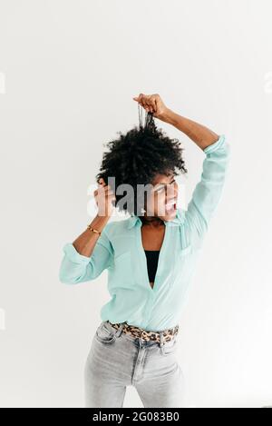 Verspielte junge afroamerikanische Hündin mit geschlossenen Augen im Trend Outfit mit Spaß berühren afro Haar auf weißem Hintergrund Stockfoto