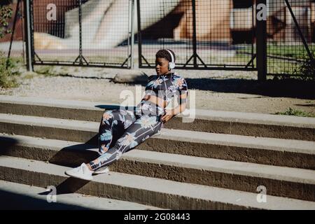 Die junge afroamerikanische Frau in farbenfroher Sportkleidung hört Musik in Kopfhörern, während sie auf einer Steintreppe im Park sitzt Stockfoto