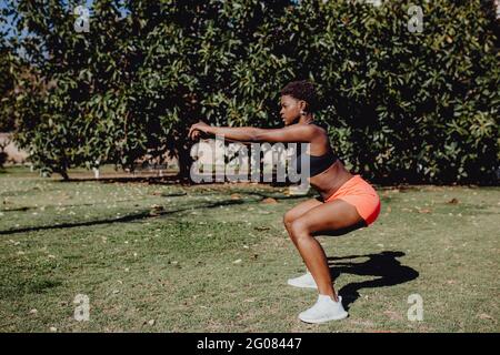Seitenansicht einer jungen afroamerikanischen Fitness-Frau, die an einem sonnigen Tag im Park hockt Stockfoto