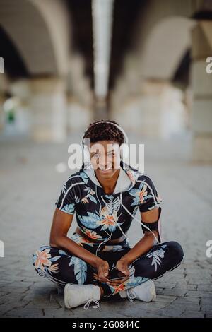 Junge afroamerikanische Fitness-Frau, die sich ausruhte und die Beine streckte, während sie Musik hörte, nachdem sie auf der Stadtstraße gelaufen war Stockfoto