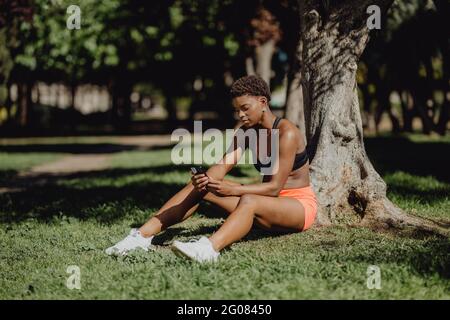 Afroamerikanische Frau in Sportkleidung hält Smartphone, während sie an sonnigen Tagen auf Gras sitzt Stockfoto