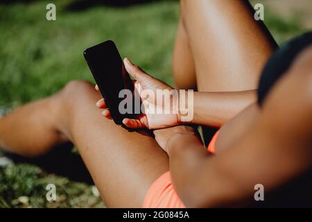 Hände einer afroamerikanischen Frau in Sportkleidung, die ihr Smartphone hält, während sie an sonnigen Tagen auf dem Gras sitzt Stockfoto