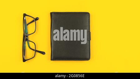Business-Schreibtisch mit schwarzem Ledernotepad und Brille, Draufsicht, gelber Tischhintergrund mit Kopierfläche Stockfoto