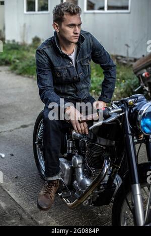 Hübscher Mann in Doppeljeansstoff auf dem Motorrad sitzend. Porträt eines hübschen jungen Mannes auf einem Vintage-Motorrad-Café-Rennfahrer. Stockfoto
