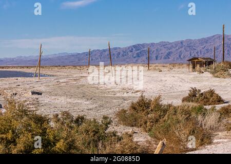 USA, CA, Salton Sea - 28. Dezember 2012: SE-Strand von Niland Marina mit ruinöser Holzhütte und elektrischen Stangen auf beigem Sand unter blauem Himmel. Mounta Stockfoto