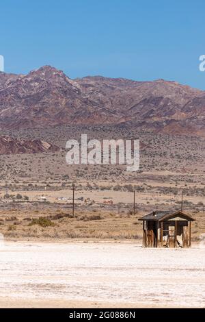 USA, CA, Salton Sea - 28. Dezember 2012: Porträt einer braunen Holzhütte auf weißem Sand vor einer hohen braunen Bergkette unter blauem Himmel. Stockfoto