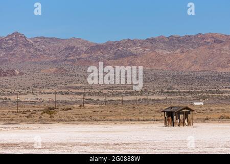 USA, CA, Salton Sea - 28. Dezember 2012: Landschaft mit brauner Holzhütte auf weißem Sand vor einer hohen braunen Bergkette unter blauem Himmel. Stockfoto
