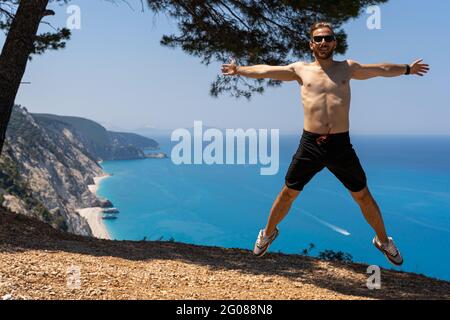 Junger Mann, der am Aussichtspunkt am Strand von Egremni springt und seine Arme und Beine ausbreitet Stockfoto