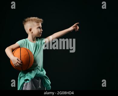 Lächelndes, blondes Kind in einem grünen T-Shirt und einer Hose, die Basketball in den Händen hält und mit dem Finger nach oben zeigt Stockfoto