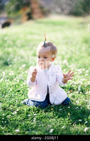 Das kleine Kleinkind mit einer Kamille hinter ihrem Ohr sitzt auf einem grünen Rasen inmitten weißer Blumen Stockfoto