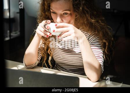 Junge kaukasische Frau, die Kaffee trinkt, während sie sich am frühen Morgen ein Video auf einem Laptop-Computer zu Hause ansieht Stockfoto