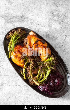 Piri- oder Peri-Hühnchen-Abendessen mit geröstetem Brokkoli und eingelegtem Rotkohl auf einem schwarzen Teller. Auf einem konkreten Hintergrund Stockfoto