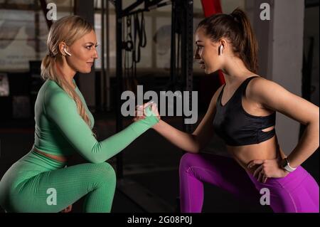 Zwei schöne junge Sportmädchen in Sportkleidung posieren im Fitnessstudio. Blonde und Brünette halten sich beim Armringen die Hände. Stockfoto