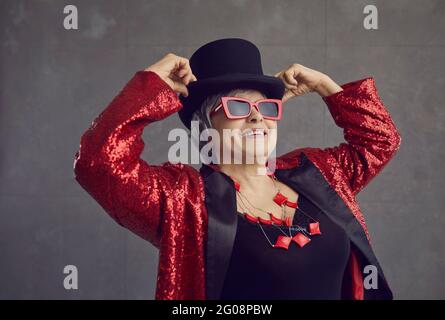 Fröhliche, schöne ältere Frau mit Zylinderhut, stilvollen Gläsern und Bling Jacket, die im Studio posiert Stockfoto