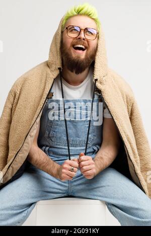 Bärtiger Mann mit modischer Jacke und Denim-Overalls, der positive Emotionen ausdrückt, lacht, optimistisch aufschaut, auf Würfel sitzt. Innen s Stockfoto