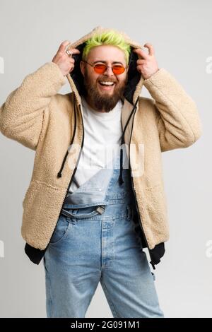 Glückliches Hipster-Modell mit Bart und hellen Haaren, Blick auf die Kamera und Lachen laut, setzen auf Kapuze, positive Emotionen auszudrücken. Studio im Innenbereich Stockfoto