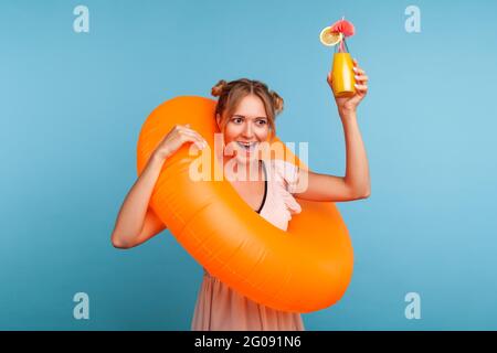Attraktive Frau mit zwei lustigen Haarbrötchen, die lächelnd zur Kamera schauen, Gummiring halten, Glas mit Saft oder Cocktail aufsteigen und vor Aufregung brüllen Stockfoto