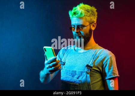 Bärtig konzentrierter Hipster-Mann, der das Handy in den Händen hält, soziale Netzwerke überprüft, neue Beiträge schreibt, hat ernsthaften Ausdruck und trägt Denim-Overal Stockfoto