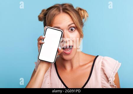Unglaubliche Werbung auf mobilen Geräten! Erstaunt schöne Frau in Kleid bedeckt Auge mit Smartphone und halten den Mund offen in Überraschung. Innen s Stockfoto