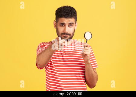 Serious selbstbewussten Mann Privatdetektiv in gestreiftem T-Shirt halten in der Hand Lupe und zeigen Finger auf Sie Blick auf Kamera, Spionage. Indo Stockfoto