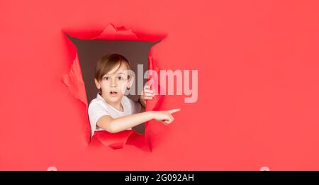 Junge in rotem Papierloch, der mit dem Finger zur Seite zeigt. Überraschender Junge, der durch ein Loch in der Papierwand schaute. Schockiert Kind macht Loch in Papier. Kinderlooks Stockfoto