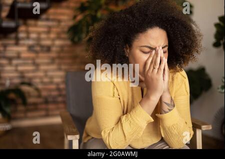 Verstörte afroamerikanische Frau sitzt auf dem Sessel und versteckt sich mit Handflächen, weint und fühlt sich depressiv, hat Probleme Stockfoto