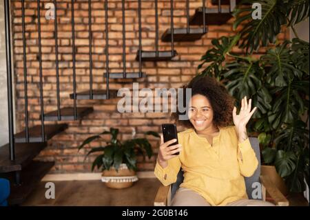 Fröhliche afroamerikanische Frau, die per Videoanruf auf dem Smartphone spricht, auf dem Sessel im gemütlichen Wohnzimmer sitzt, Hallo in die Webcam winkt, glücklich, Freunde oder Familie auf der Ferne zu treffen Stockfoto