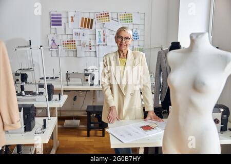 Reife Frau mit Brille steht am Tisch mit Kleidungszeichnungen in der Nähwerkstatt Stockfoto