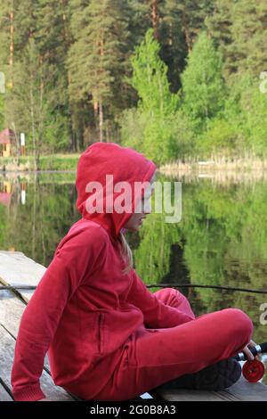 Ein blondes Teenager-Mädchen in einem roten Anzug sitzt auf einer Holzbrücke an einem Waldsee und hält eine Angelrute in ihren Händen. Portrait eines blonden Mädchens, das angeln soll Stockfoto