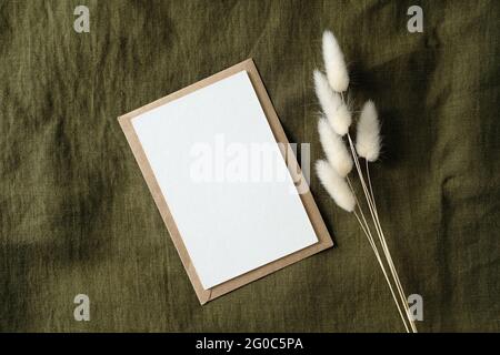 Weiße Einladungskarte Mockup und getrocknete Blumen auf grünem Stoff Draufsicht. Einladungskarte für Vintage-Hochzeiten. Stockfoto