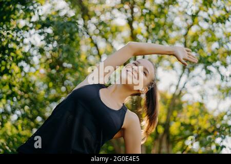 Sportliche Frau macht Aufwärmen und Stretching-Übungen lehnt sich zur Seite hat Inhalt Gesichtsausdruck in aktiven tragen Posen vor Natur Hintergrund gekleidet Stockfoto