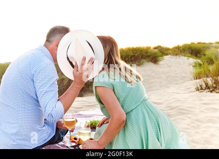 Anonymer Mann und Frau mit Weinläsern, die während eines romantischen Picknicks mit Hut am Strand Toast auf Obst anbieten Stockfoto