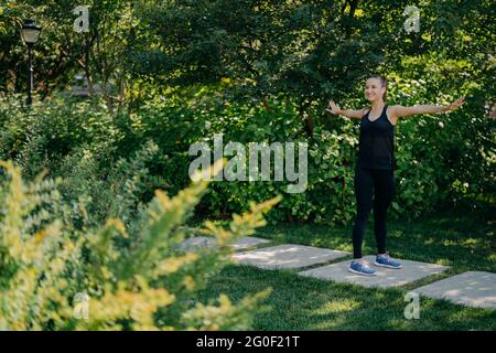 Erfreut Frau streckt Arme seitwärts erwärmt sich vor dem morgendlichen Training trägt schwarze Sportklotzen und Turnschuhe hört Musik in Kopfhörer genießt die Natur Stockfoto
