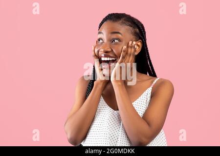 Die hübsche afroamerikanische Frau drückt ihre Aufregung aus, berührt überrascht ihr Gesicht und blickt auf einen rosa Hintergrund Stockfoto