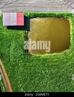 Luftaufnahme eines Schuppen hinter einem kleinen künstlichen Fischteich auf einem Weg durch eine grüne Wiese neben einem Feld, abstrakter Effekt Stockfoto