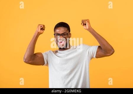 Emotionaler afroamerikanischer Mann, der WOW in Aufregung schreit und die Arme über seinen Kopf über gelben Hintergrund hebt Stockfoto