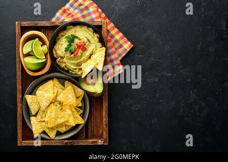 Guacamole. Traditionelle lateinamerikanische mexikanische Dip-Sauce in einer schwarzen Schüssel mit Avocado und Zutaten und Maisnachos. Avocadoverbreitung. Draufsicht. Copyspa Stockfoto