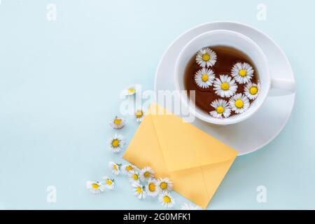 Eine Tasse Kamillentee, ein Umschlag und Gänseblümchen auf blauem Hintergrund. Draufsicht, flach liegend, Kopierbereich. Stockfoto