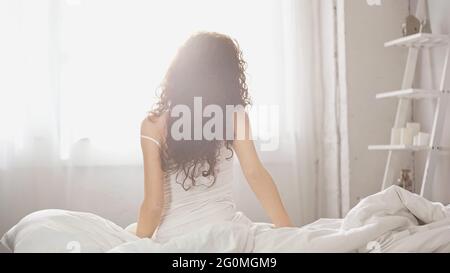 Rückansicht einer jungen lockigen Frau, die auf dem Bett sitzt Stockfoto