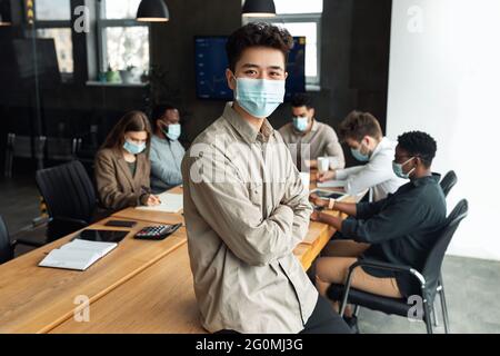 Junger asiatischer Geschäftsmann in Maske sitzt auf dem Schreibtisch und posiert Stockfoto