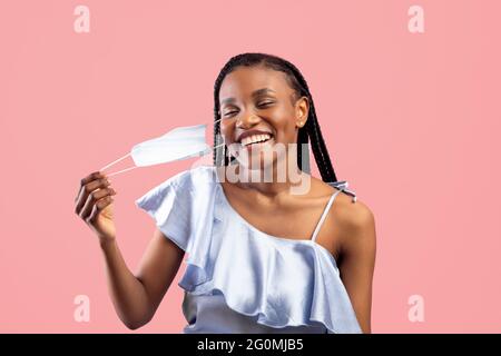 Coronavirus ist vorbei. Glückliche schwarze Frau mit afro Bunches, die ihre Gesichtsmaske auf pinkem Studiohintergrund ausziehen Stockfoto