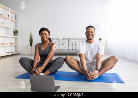 Inländische Schulungen. Junges schwarzes Paar, das mit einem Online-Lehrer am Laptop Yoga praktiziert und zu Hause Schmetterling Asana macht Stockfoto